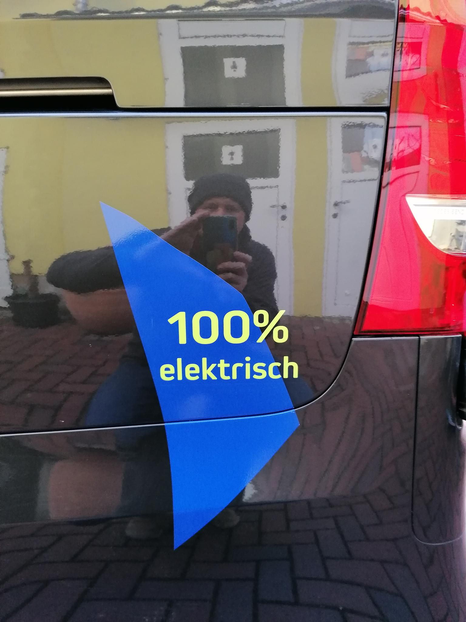 Philipp Wedekind spiegelt sich beim fotographieren im schwarzen Lack eines Autos mit der Aufschrift 100% elektrisch